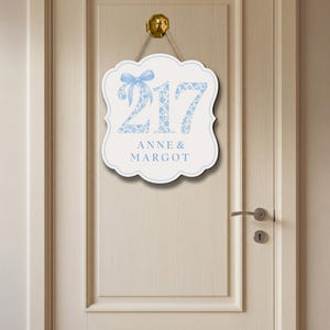 May include: A white scalloped wall sign featuring the number 217 in a light blue floral pattern, accented by a light blue bow graphic. Below the number, the text ANNE & MARGOT is printed in light blue. The sign hangs from a gold-colored hook.