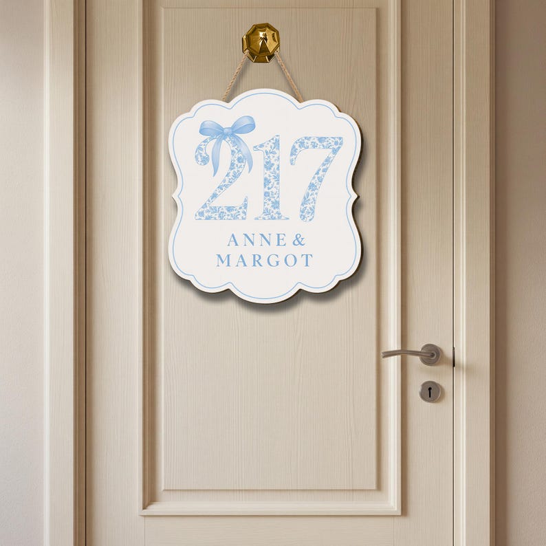 May include: A white scalloped wall sign featuring the number 217 in a light blue floral pattern, accented by a light blue bow graphic. Below the number, the text ANNE & MARGOT is printed in light blue. The sign hangs from a gold-colored hook.