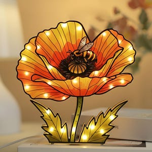 May include: A decorative poppy flower night light with a bee on the flower. The flower is orange and yellow with a black outline and small lights. The base has green leaves and the text "The art of libraries".
