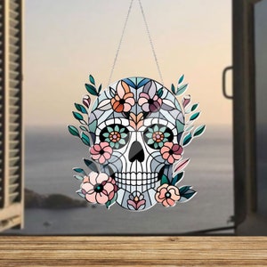 May include: A colorful stained glass sugar skull with floral accents hanging from a silver chain. The skull is blue, white, and black with pink and green flowers.