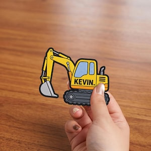 Op de afbeelding: Een gele en zwarte graafmachine patch met de naam KEVIN in zwarte hoofdletters op de zijkant van de cabine. Het ontwerp van het bouwvoertuig bevat een grijze bak, een lichtblauw raam en zwarte rupsbanden met een dikke zwarte rand.