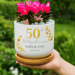 May include: A white ceramic planter with a succulent plant featuring bright pink flowers. The planter is decorated with gold text that reads "50th Wedding Anniversary" and the names "JOHN & JANE" with the date "09.18.1974".
