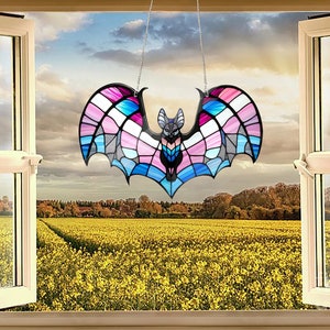 May include: A stained glass bat suncatcher with a black frame and a colorful design of pink, blue, and white. The bat is hanging in front of an open window overlooking a field of yellow flowers.