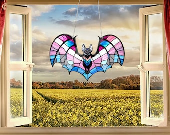 May include: A stained glass bat suncatcher with a black frame and a colorful design of pink, blue, and white. The bat is hanging in front of an open window overlooking a field of yellow flowers.