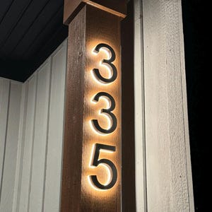 May include: Illuminated house numbers 335 mounted on a wooden post. The numbers are black with a warm, glowing light outlining each digit. The post is a dark brown color, and the background includes white siding.