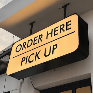 May include: A rectangular, double-sided, illuminated sign mounted to a building exterior. The sign features a warm yellow background with black text that reads ORDER HERE on the top line and PICK UP on the bottom line, separated by a line.