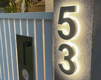 Puede incluir: Un pilar gris oscuro texturizado con los números de casa 53 iluminados en una fuente sans-serif negrita. Los números están retroiluminados con un brillo blanco cálido, creando un efecto de halo. Un buzón de metal vertical está fijado al lado.