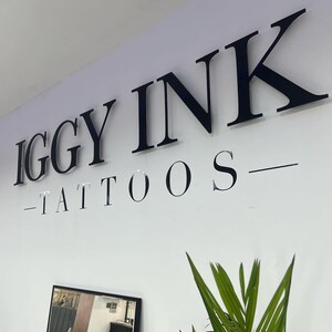 May include: Black and white sign with the text "IGGY INK - TATTOOS" on a white wall.