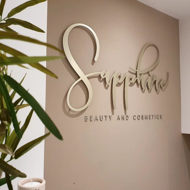 May include: A gold-colored sign with the word "Sapphire" written in cursive script. The sign also includes the words "Beauty and Cosmetics" in a smaller font.