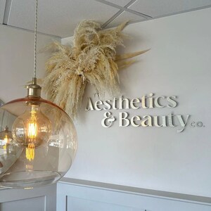 May include: A white wall with a sign that reads "The Aesthetics & Beauty Co." in gold letters. A large bouquet of dried pampas grass hangs above the sign. A gold globe light fixture hangs from the ceiling.