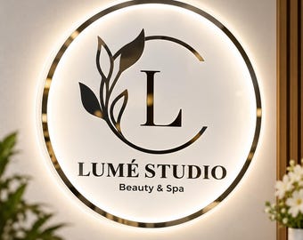 May include: A circular wall sign with a white face and a metallic gold rim featuring backlighting. The sign displays the letter L, a black leaf graphic, and the black text LUMÉ STUDIO Beauty & Spa in a clean, sans-serif font.