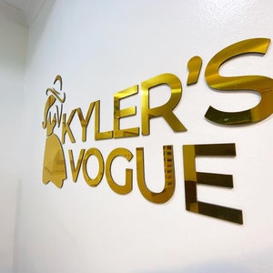 May include: Gold-colored sign with the text "KYLER'S VOGUE" and a stylized female figure wearing a hat and dress.