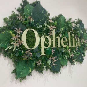 May include: A green and white sign with the word "Ophelia" in gold letters. The sign is surrounded by artificial greenery, including leaves and flowers.