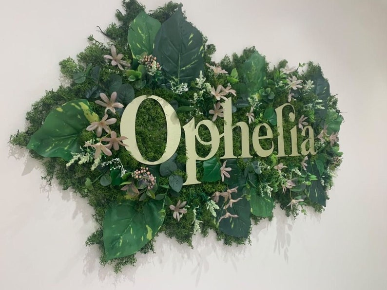 May include: A green and white sign with the word "Ophelia" in gold letters. The sign is surrounded by artificial greenery, including leaves and flowers.