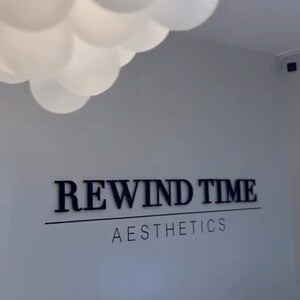 May include: A white wall with a black sign that reads "REWIND TIME AESTHETICS".