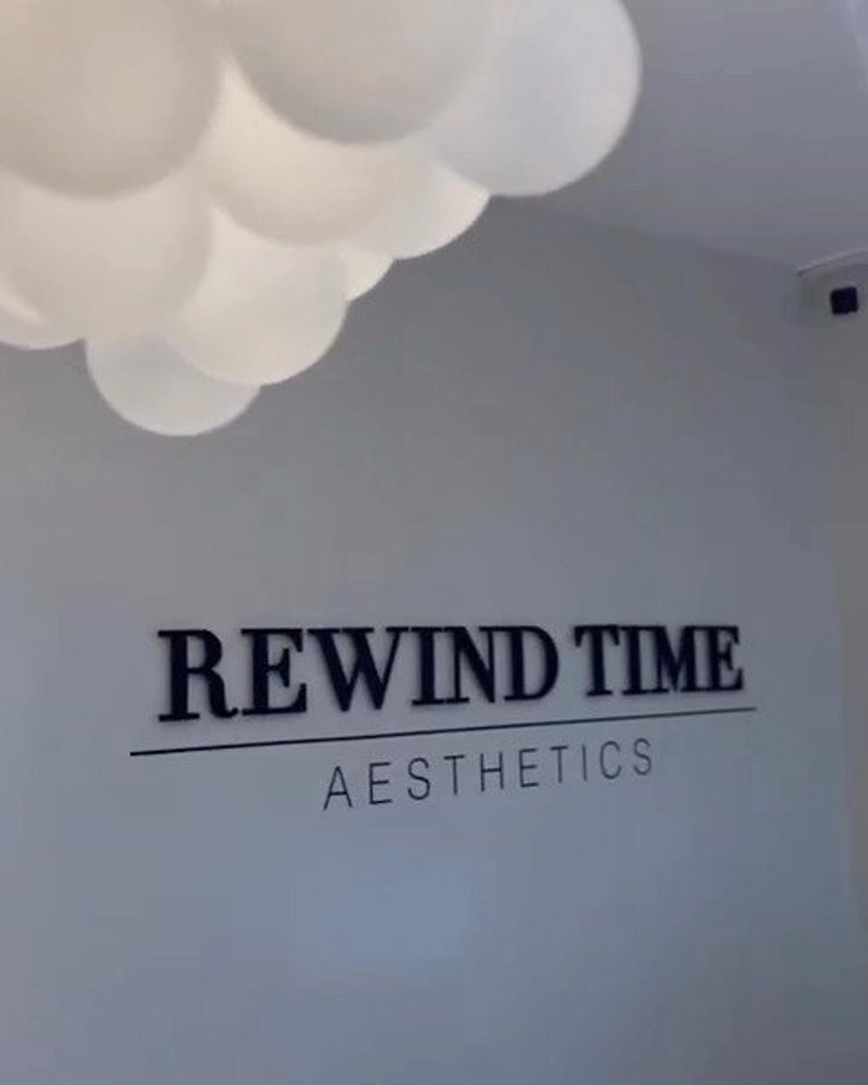 May include: A white wall with a black sign that reads "REWIND TIME AESTHETICS".