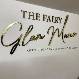 May include: Gold lettering on a white wall reads "The Fairy Glam Mama Aesthetics SPMU & Training Academy".