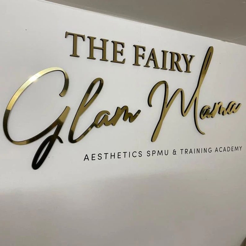 May include: Gold lettering on a white wall reads "The Fairy Glam Mama Aesthetics SPMU & Training Academy".