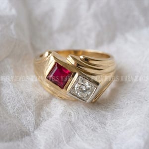 May include: A gold ring featuring a square-cut red ruby and a square-cut clear diamond set side-by-side in a bypass-style band. The band has a polished finish with textured, sweeping lines along the sides. The text MIARA JEWELS is visible.