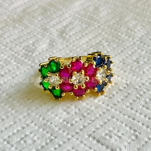 Puede incluir: Un anillo dorado con un diseño floral, adornado con un grupo de piedras preciosas de colores. El anillo muestra una vibrante disposición de piedras rojas, verdes, azules y blancas, contrastando con la banda dorada. El anillo está sobre una superficie blanca.