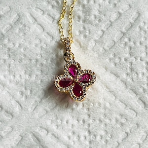 Könnte beinhalten: Eine goldene Kettenkette mit einem Anhänger in Form eines roten und weißen Schmetterlings. Der Schmetterling hat drei rote Edelsteine und ist von weißen Edelsteinen umgeben.