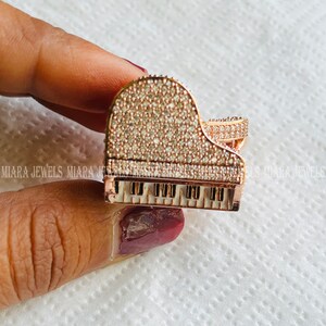 May include: A gold-tone piano pendant with a white enamel keyboard and a clear crystal design. The pendant is set with clear crystals and has a detailed design.