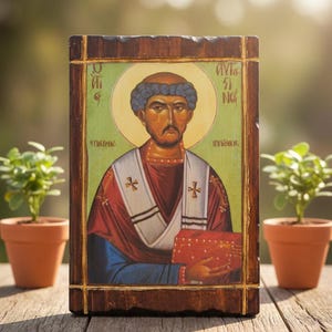 Op de afbeelding: Een rechthoekig houten icoon met een geschilderd portret van een heilige met een aureool, gekleed in rode en blauwe gewaden met een witte stola. De figuur houdt een rood boek vast. Het icoon heeft een donkerbruine lijst met gouden lijnen.