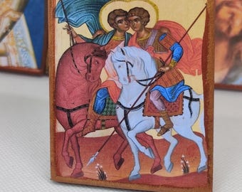 May include: A rectangular wooden icon depicting two figures on horseback, with text in Greek. The riders are in armor, holding spears, and are accompanied by a third figure. The horses are red and white, set against a warm, earthy background.
