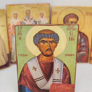 Op de afbeelding: Een houten icoon met een religieuze figuur met een halo, gekleed in een rood en wit gewaad met een blauw onderkleed. Het icoon heeft Griekse tekst en is omringd door andere soortgelijke iconen.