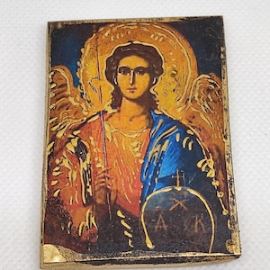 Op de afbeelding: Een klein, rechthoekig houten icoon met een gouden lijst die een geschilderd beeld van een engel met vleugels afbeeldt. De engel houdt een staf vast en heeft een halo. De achtergrond is donkerblauw met gouden accenten. Het icoon heeft de letters "A", "K" en een kruis onderaan.
