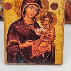 以下が含まれることがあります： 女性が子供を抱いている宗教的なアイコン。どちらにも光輪があります。女性は濃いマルーン色のローブを着ており、子供は金の衣服を着ています。背景は金で、テキストが入っています。アイコンは木のブロックにあります。