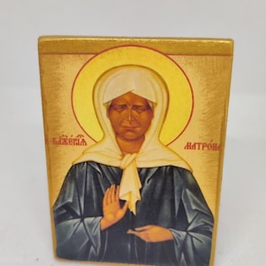 May include: A wooden icon featuring a religious figure with a golden halo and background. The figure wears a white head covering and dark robes. The icon has text in a foreign language.