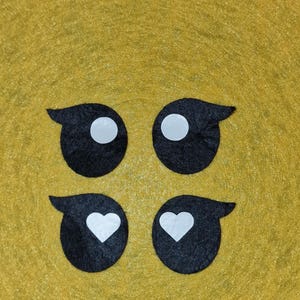 May include: Four black felt eye shapes with white circular pupils and a curved top detail are arranged on a yellow background. Two of the eye shapes have white heart-shaped pupils instead of circles.