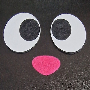 Op de afbeelding: Een zwart vilt handwerk met twee grote witte glimmende ogen en een roze vilt neus.