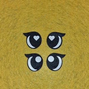 Könnte beinhalten: Vier schwarz-weiße Cartoon-Augenausschnitte mit einem Herz in den oberen Augen und einem weißen Kreis in den unteren Augen. Die Augen sind in einem quadratischen Muster auf einem gelben Hintergrund angeordnet.