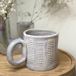 May include: A lavender ceramic mug with a distinctive textured design. The mug has a large, rounded handle and is presented on a wooden surface. The design incorporates squares and lines, creating a tactile surface. The mug is suitable for hot beverages.