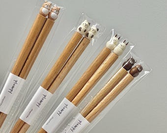 Puede incluir: Cuatro pares de palillos de madera con tapas en forma de animales. Las tapas son un perro marrón y blanco, un gato blanco, un conejo blanco y un oso marrón. Los palillos están empaquetados en plástico transparente.