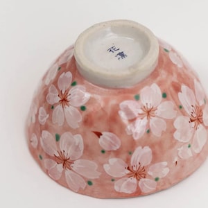 Puede incluir: Un cuenco de cerámica rosa con un diseño de flores de cerezo blancas. La parte inferior del cuenco tiene los caracteres japoneses "花 窯" en azul.