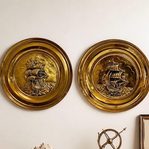 Pode incluir: Duas placas de parede redondas douradas com desenhos de navios à vela em relevo. As placas têm uma borda elevada e uma representação detalhada de um navio no mar. Estão montadas numa parede branca, criando um tema náutico.