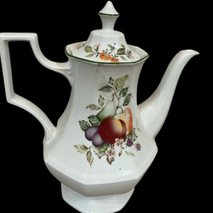 Könnte beinhalten: Weiße Kaffeekanne aus Keramik mit einem grünen und braunen Blumendesign mit Früchten und Beeren. Die Kanne hat einen Deckel mit einer spitzen Oberseite.