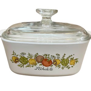 Puede incluir: Plato de cerámica blanco para hornear con tapa de vidrio. El plato tiene un diseño floral con verduras y el texto "L'Echalote".