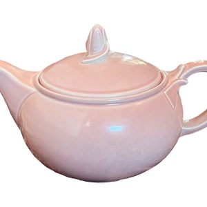 May include: A pink ceramic teapot with a lid and a floral-shaped knob. The teapot has a rounded body and a spout on one side and a handle on the other.