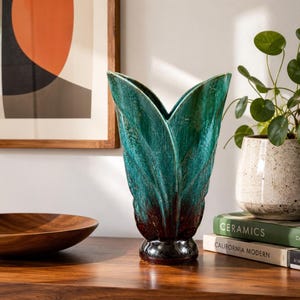 Peut inclure: Un vase en céramique avec une silhouette en forme de feuille, présentant un émail vert sarcelle profond qui se transforme en une base brun foncé. Le vase possède un bord évasé à plusieurs pointes et un pied circulaire, avec une finition texturée.