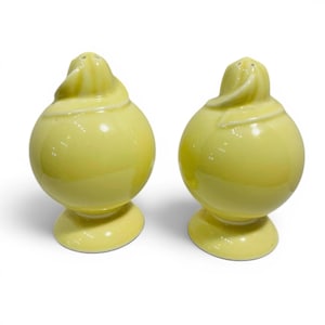 May include: A pair of yellow ceramic salt and pepper shakers. Each shaker has a bulbous body and a swirl design on top. The shakers sit on a small, round base. The color is a pale, sunny yellow.