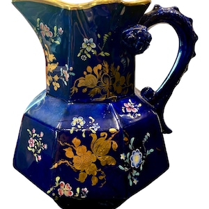 May include: A cobalt blue ceramic pitcher with a gold rim and floral designs. The pitcher has a unique handle with a sculpted design.