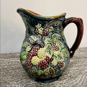 May include: A blue and green ceramic jug with a brown handle. The jug is decorated with a raised design of berries and leaves.
