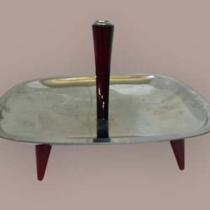 Könnte beinhalten: Ein silbernes Serviertablett mit einem roten Griff und drei roten Beinen. Das Tablett ist quadratisch und hat einen leicht erhöhten Rand.