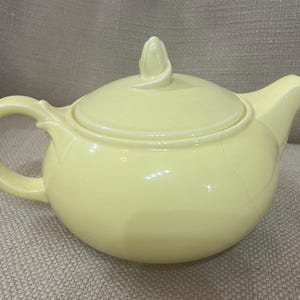 Pode incluir: Uma chaleira de cerâmica de cor amarelo claro com um corpo arredondado e uma única alça. A chaleira tem uma tampa com um botão decorativo em forma de folha.