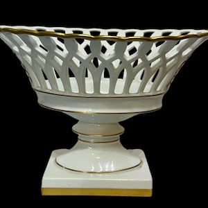 Op de afbeelding: Een witte porseleinen mand met een traliewerkontwerp, geaccentueerd met gouden randen. De mand staat op een voetstuk met een vierkante voet, eveneens afgewerkt met goud. Het elegante stuk is geschikt voor decoratief gebruik.