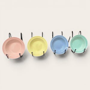 Puede incluir: Cuatro platos de cerámica de colores pastel, rosa, amarillo, azul y verde, se exhiben en una pared blanca. Cada plato está sujeto por un gancho de metal negro. Los platos son redondos con un centro ligeramente hundido.